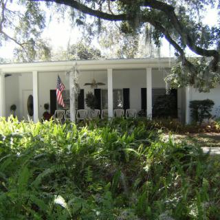 A white house with a porch and columns surrounded by lush greenery and trees.