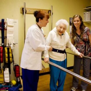 An elderly person receiving support from healthcare professionals during a physical therapy session.