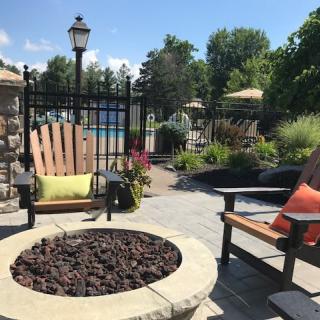 A cozy outdoor patio with a fire pit, surrounded by chairs and lush greenery, near a pool on a sunny day.