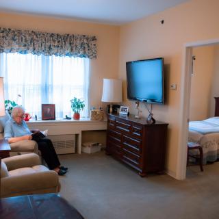 A cozy living room with a person sitting in an armchair, watching TV, and a view into the adjacent bedroom.