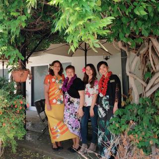 A group of women standing together in a garden, wearing colorful traditional clothing and lei necklaces.