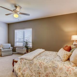 A cozy bedroom with a floral bedspread, a comfortable chair by the window, and a ceiling fan overhead.