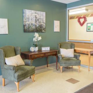 A cozy waiting area featuring two green upholstered chairs, a decorative table, and calming artwork on the walls.