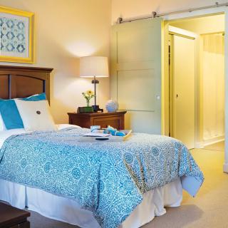A cozy bedroom with a blue-patterned bedspread, a nightstand with a lamp, and an open sliding door.