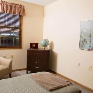 A cozy bedroom with a dresser, globe, chair, and window with blinds.