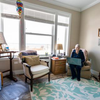 An elderly person sitting in an armchair in a cozy living room with large windows.