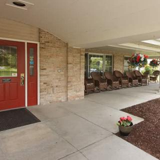 A welcoming building entrance with a red door, lined with rocking chairs and flower pots along the porch.