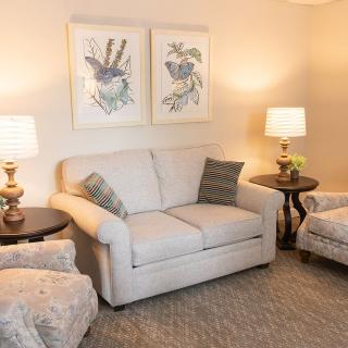 A cozy living room with a beige sofa and matching armchairs, accompanied by side tables with lamps and floral artwork on the wall.