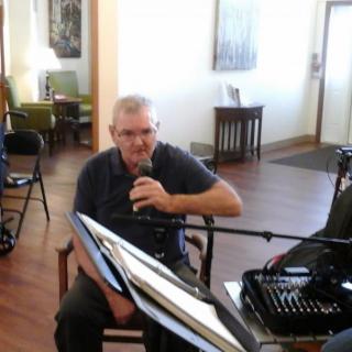 A man sitting indoors holding a microphone, with a piano nearby.