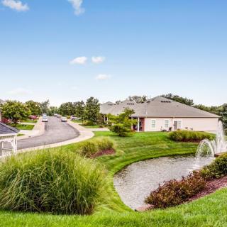 A peaceful suburban neighborhood with well-maintained houses, lush greenery, and decorative fountains along the road.