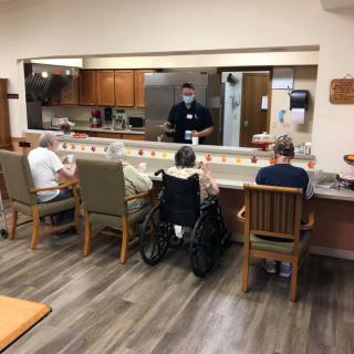 Residents gather in a care facility dining area, interacting with staff.