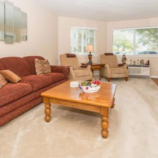 A cozy living room featuring a brown sofa, two armchairs, and a wooden coffee table on a light carpet, with large windows providing natural light.