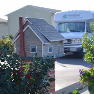 A miniature house mailbox sits beside a driveway with an RV parked in the background.