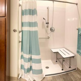 An accessible bathroom shower with a blue and white striped curtain, equipped with a shower bench, handheld showerhead, and safety grab bar.