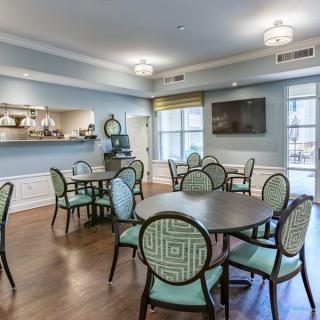 A well-lit dining area featuring several round tables and cushioned chairs, with a television mounted on the wall and large windows providing natural light.