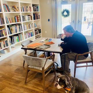 A man sits thoughtfully at a table surrounded by books, with a dog resting nearby.