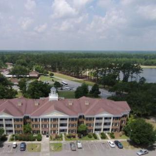 Aerial view of a residential building with a parking lot, surrounded by trees and a nearby lake under a cloudy sky.
