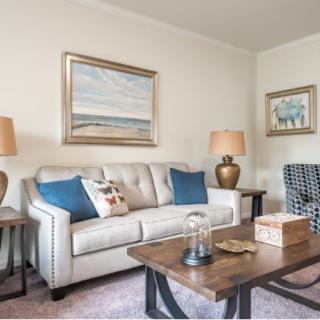 A cozy living room featuring a beige sofa with blue and patterned cushions, two side tables with matching lamps, a wooden coffee table, and decorative wall art.