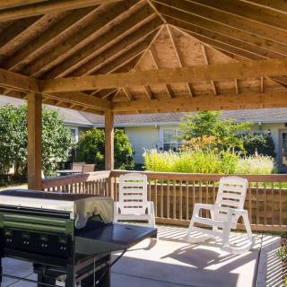 A wooden gazebo with patio chairs and a grill in a garden setting.
