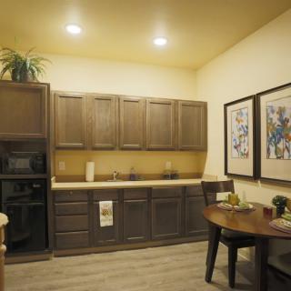 A modern kitchen interior featuring wood cabinets, overhead lighting, and a small dining area with framed artwork on the walls.