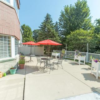 A sunny patio with tables, chairs, and red umbrellas, surrounded by trees.