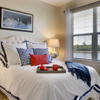 A cozy bedroom with elegant decor, featuring a neatly made bed with decorative pillows and a tray, illuminated by a lamp beside the window.