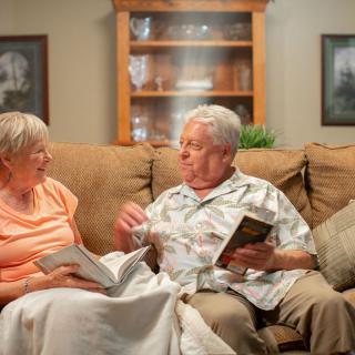An elderly couple sits on a couch in their living room, engaged in conversation while holding books.