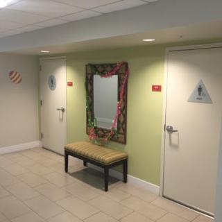 A hallway featuring restroom signs, a mirror, and a bench.