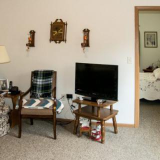 A cozy living room with a TV, chair draped with a plaid blanket, and a side table in front of a doorway leading to another room.