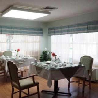 A well-lit dining room with neatly set tables and chairs, adorned with simple decor and surrounded by large windows.