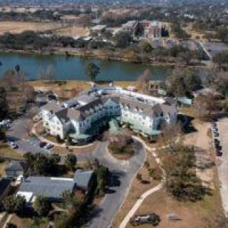 Aerial view of a building complex near a body of water, surrounded by trees and roads.