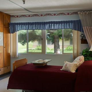 A cozy bedroom featuring a red bedspread, large windows with blue curtains, and warm wooden furniture.
