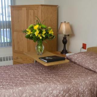 A cozy bedroom with a neatly made bed, a small table, a lamp, and a vase of flowers.