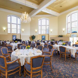 A spacious dining hall with elegantly set tables and comfortable chairs, illuminated by a grand chandelier and large windows.
