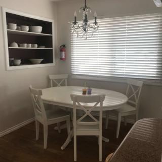 A cozy dining area with a round white table, four matching chairs, and a chandelier overhead. Natural light streams in through the window blinds, highlighting the neatly arranged dishes on open shelves.