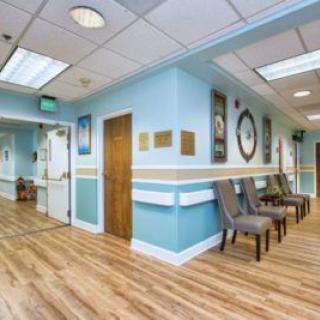 A well-lit waiting room with light blue walls and wooden flooring, featuring a row of chairs and a large clock on the wall.
