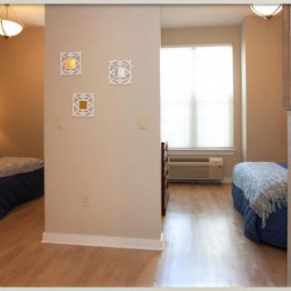 A cozy bedroom with two beds separated by a partition wall, featuring wood flooring and natural sunlight.