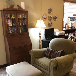 A cozy living room corner with an armchair, ottoman, and a wooden bookshelf, accompanied by a floor lamp and decorative plates on the wall.
