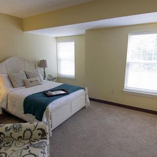 A cozy and well-lit bedroom featuring a comfortable bed with a teal blanket and decorative pillows, illuminated by natural light from the large windows.
