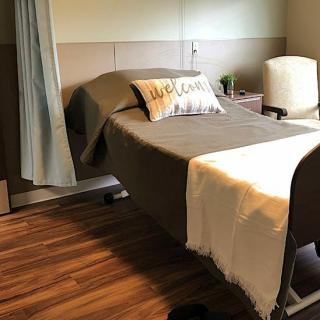 A room with a hospital bed, pillow, blanket, and a chair on a wooden floor.