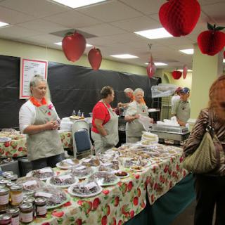 A bustling community market featuring various jams, jellies, and local treats, surrounded by festive strawberry decorations.