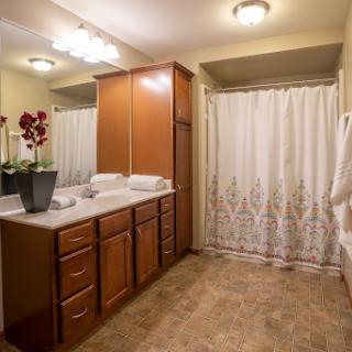 A cozy bathroom featuring wooden storage cabinets, a well-lit vanity area, and elegant touches like decorative plants and a shower curtain.