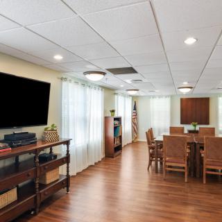 A well-lit conference room with wooden flooring, featuring a large television, a table surrounded by chairs, and decorative elements.