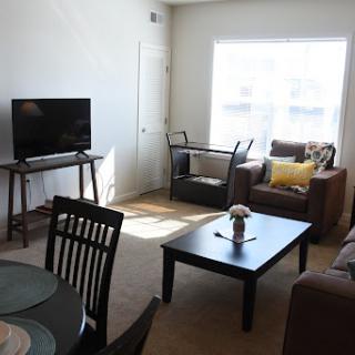A cozy living room featuring a television, comfortable seating, and a dining area with natural light streaming in.