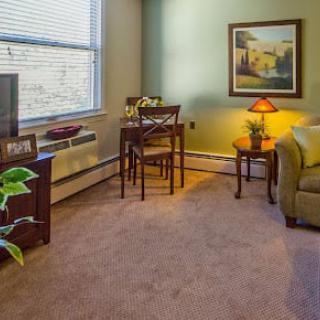 A cozy living room featuring a sofa, table with chairs, lamp, artwork on the wall, and a potted plant by the window.
