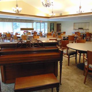 A spacious dining room with a piano, multiple tables and chairs, large windows, and chandeliers.