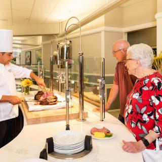 A chef serves freshly carved meat at a buffet while two customers watch eagerly.