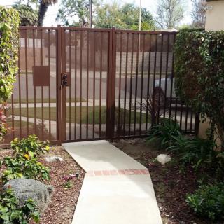 A brown iron gate with a concrete pathway leading to it, surrounded by greenery.