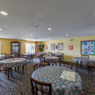 A brightly lit dining room with several round tables covered in patterned tablecloths, surrounded by chairs and decorated with wall art.