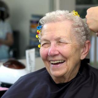 An elderly woman smiling while getting her hair done at a salon.